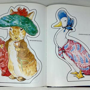 May include: A coloring book page featuring a cutout of a rabbit wearing a green hat and a duck wearing a blue hat. The rabbit is holding a carrot and the duck is wearing a red and blue patterned dress. The text "Benjamin Bunny" is printed next to the rabbit and "Jemima Puddle-Duck" is printed next to the duck.
