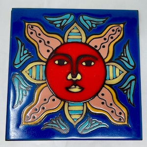 Puede incluir: Un azulejo de cerámica azul con una cara de sol roja con rayos amarillos, azules y rosas. El sol tiene una nariz amarilla, ojos negros y una boca amarilla.