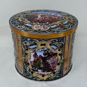 May include: A round, vintage tin with a gold and blue design. The tin features images of a European town and decorative floral elements. The lid and sides have detailed artwork, showcasing a classic, antique aesthetic.