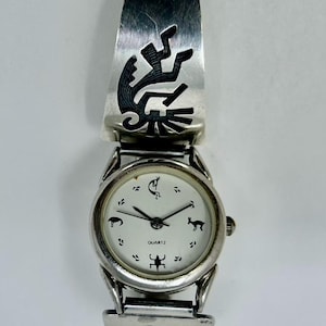 May include: A silver watch with a white face and black markings. The watch band is a cuff style with a Native American design.