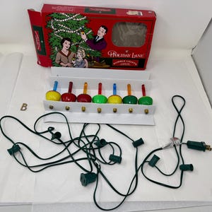 Könnte beinhalten: Ein Set Vintage Holiday Lane Blasenlichter in einer roten Box. Die Lichter befinden sich in einem weißen Halter mit einer Vielzahl von farbigen Glühbirnen, darunter Gelb, Rot und Grün. Die Box zeigt eine Familie, die einen Weihnachtsbaum schmückt.