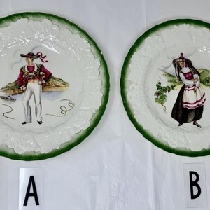 May include: Two decorative plates with green rims and white, textured edges. One plate features a man in traditional attire, the other a woman. Both plates have colorful illustrations of people in traditional clothing.
