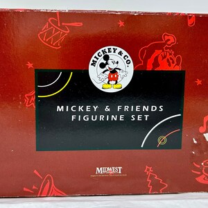 May include: Red box with a Mickey Mouse and Friends Figurine Set. The box features a Mickey Mouse logo with Mickey Mouse in a red and yellow outfit. The box also features a red and white candy cane, a red and white drum, a red and white train, a red and white Christmas tree, and a red and white musical note.
