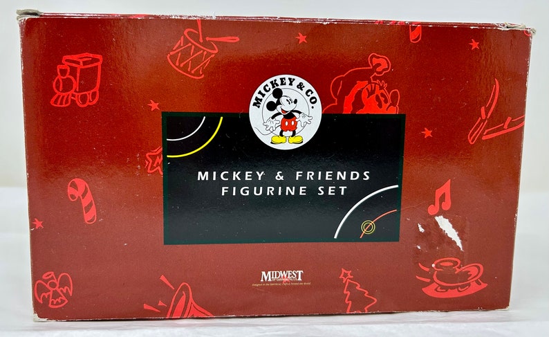 May include: Red box with a Mickey Mouse and Friends Figurine Set. The box features a Mickey Mouse logo with Mickey Mouse in a red and yellow outfit. The box also features a red and white candy cane, a red and white drum, a red and white train, a red and white Christmas tree, and a red and white musical note.