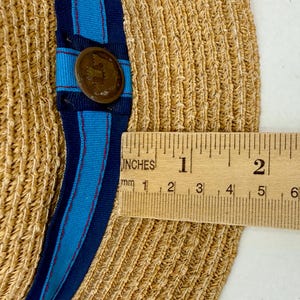 May include: A tan woven straw hat featuring a blue and navy striped ribbon band with a circular bronze-toned metal button. A wooden ruler is placed across the brim, showing a measurement of approximately 2 inches from the edge to the ribbon band.