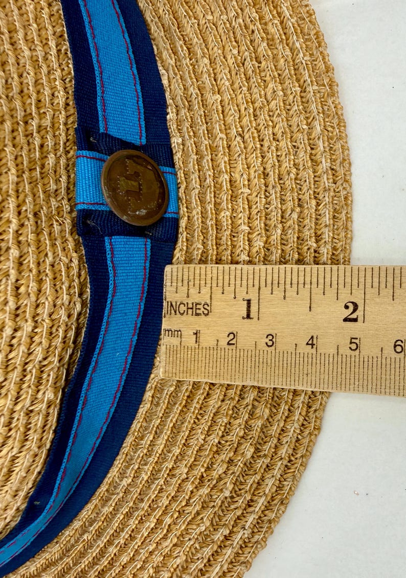 May include: A tan woven straw hat featuring a blue and navy striped ribbon band with a circular bronze-toned metal button. A wooden ruler is placed across the brim, showing a measurement of approximately 2 inches from the edge to the ribbon band.