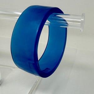 Może przedstawiać: Żywa niebieska bransoletka typu bangle. Bransoletka to solidny, gruby pas z półprzezroczystego materiału. Bransoletka jest prezentowana na przezroczystym, akrylowym stojaku, podkreślając jej gładkie, zaokrąglone krawędzie i odważny kolor. Prosty, nowoczesny dodatek.