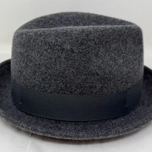 Pode incluir: Um chap&eacute;u fedora de feltro cinza-carv&atilde;o escuro com uma coroa estruturada com um vinco central e uma frente pin&ccedil;ada. O chap&eacute;u &eacute; acentuado com uma fita de gorgor&atilde;o preta lisa e s&oacute;lida que circunda a base da coroa acima da aba.