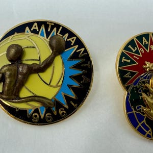 May include: Two vintage enamel badges. One depicts a volleyball player with the text "ATLANTA 1996". The other badge has a globe design with the text "ATLANTA 1996" and a starburst.