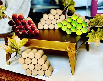 Op de afbeelding: Een gouden metalen serveerblad toont drie decoratieve druiventrossen gemaakt van houten cilinders. Eén tros is dieprood, een andere is natuurlijk hout en de derde is groen. Kunstmatige herfstbladeren en extra houten druiventrossen zijn ook aanwezig.