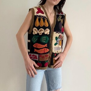 May include: A black, sleeveless, knitted vest with a V-neck, featuring colorful food-themed designs. The vest includes images of mushrooms, fish, and other produce. The vest is worn with light blue jeans.