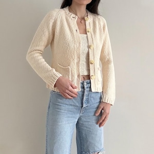 May include: A cream-colored cardigan with a textured knit, featuring round buttons and two front pockets. Worn with a matching crop top and light blue ripped jeans. The outfit is a casual, vintage-inspired ensemble.