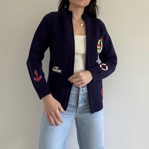 May include: Navy blue zip-up cardigan with nautical-themed embroidered patches. Features an anchor, boat, life preserver, and other maritime designs. The cardigan is worn with light blue ripped jeans.