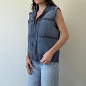 May include: A blue denim vest with a zip-up front, cable knit detailing, and a collared neckline. The vest is sleeveless and has a ribbed knit hem. The model is wearing light wash ripped jeans.