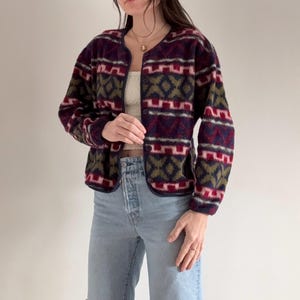 May include: A patterned fleece jacket in shades of burgundy, olive green, and white. The jacket features a geometric design and a rounded neckline. The jacket is worn with light blue ripped jeans.