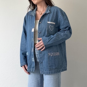 May include: A light blue denim jacket with a button-up front, featuring a heart patch on the chest pocket and floral patches. The jacket is paired with light wash jeans with distressed knees, creating a casual, vintage aesthetic.