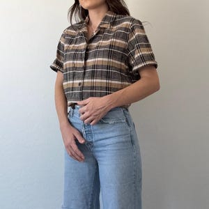 May include: A short-sleeved, collared shirt with a plaid pattern in shades of brown, tan, and black. The shirt is paired with light blue denim jeans with distressed knee details. The outfit is casual and stylish.