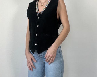 May include: A black velvet vest with a colorful beaded trim and button closure. The vest is worn over a pair of light blue jeans with distressed details.