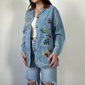 May include: A light blue denim jacket with a brown leather collar and colorful leaf and flower embroidery. The jacket has a button-up front and two pockets.