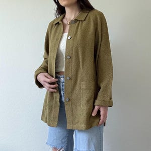 May include: A green tweed jacket with a button-up front and pockets. The jacket is worn over a white tank top and blue jeans with distressed details.