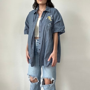 May include: Blue and white checkered short-sleeve button-down shirt with a cartoon character graphic on the chest. The shirt is worn over a white top and paired with light blue ripped jeans.