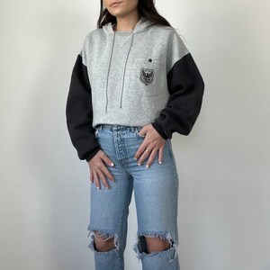 May include: A gray and black hooded sweatshirt with a small embroidered design on the front pocket. The sweatshirt has a relaxed fit and is made of a soft, comfortable fabric.