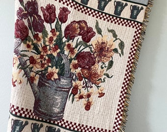 May include: A cream-colored woven throw blanket with a floral pattern featuring red, yellow, and green flowers in a metal watering can. The blanket has a red and white checkered border and a blue and green border with a repeating pattern of watering cans.