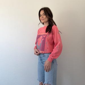 May include: A coral-coloured sweatshirt featuring a floral design in pink and purple. The long-sleeved top is worn with light-wash, ripped-knee jeans. The floral design is centered on the chest.