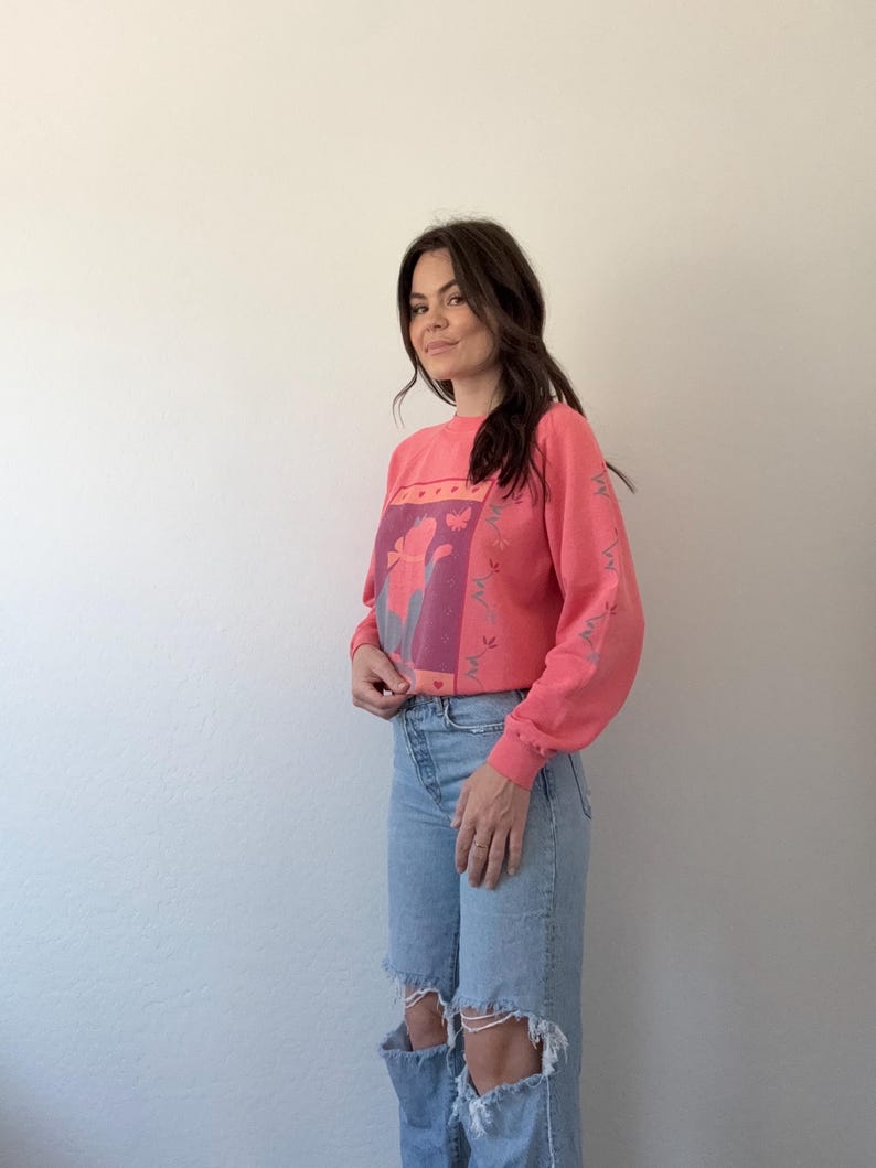 May include: A coral-coloured sweatshirt featuring a floral design in pink and purple. The long-sleeved top is worn with light-wash, ripped-knee jeans. The floral design is centered on the chest.