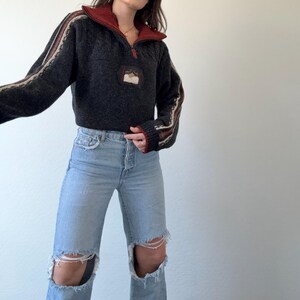 Puede incluir: Un su&eacute;ter gris oscuro con cuello rojo y cremallera de cuarto. Las mangas tienen detalles a rayas beige y marr&oacute;n. El su&eacute;ter se combina con vaqueros rotos de color azul claro.