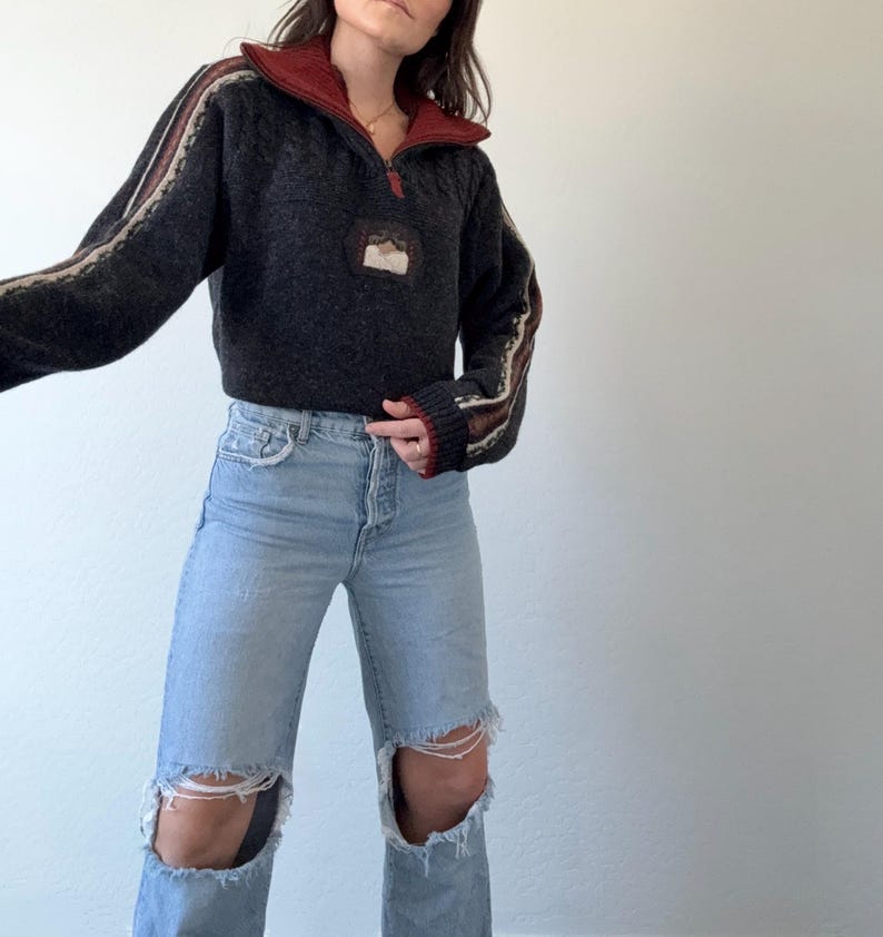 Puede incluir: Un su&eacute;ter gris oscuro con cuello rojo y cremallera de cuarto. Las mangas tienen detalles a rayas beige y marr&oacute;n. El su&eacute;ter se combina con vaqueros rotos de color azul claro.