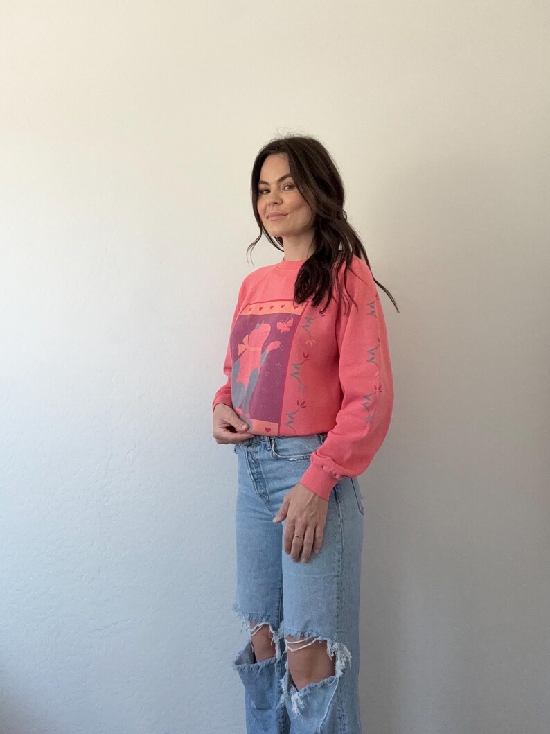 May include: A coral sweatshirt with a floral design in purple and pink. The long-sleeved top has a crew neck and a relaxed fit. The model is wearing light blue ripped jeans.