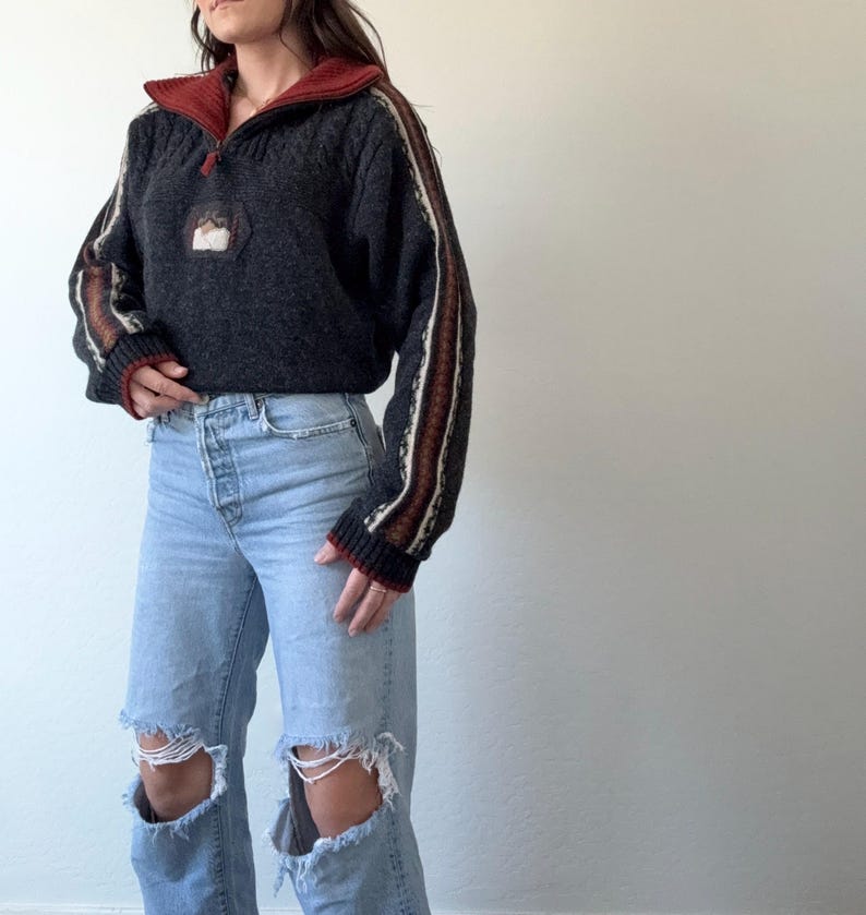 Puede incluir: Un su&eacute;ter gris oscuro con cuello y pu&ntilde;os rojos, con cierre de media cremallera y un peque&ntilde;o parche bordado. Las mangas tienen detalles a rayas en blanco, marr&oacute;n y rojo. El su&eacute;ter se combina con vaqueros rotos de color azul claro.