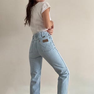 May include: Light blue Wrangler jeans with a high waist and a brown leather patch on the back pocket. The jeans are paired with a white, short-sleeved, open-knit top. The jeans have a classic, straight-leg fit.