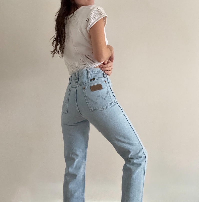 May include: Light blue Wrangler jeans with a high waist and a brown leather patch on the back pocket. The jeans are paired with a white, short-sleeved, open-knit top. The jeans have a classic, straight-leg fit.