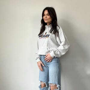 May include: A gray hooded sweatshirt with a Mickey Mouse graphic and the text "Mickey" and "Disney World" printed on the front. The model is wearing the sweatshirt with light blue ripped jeans.