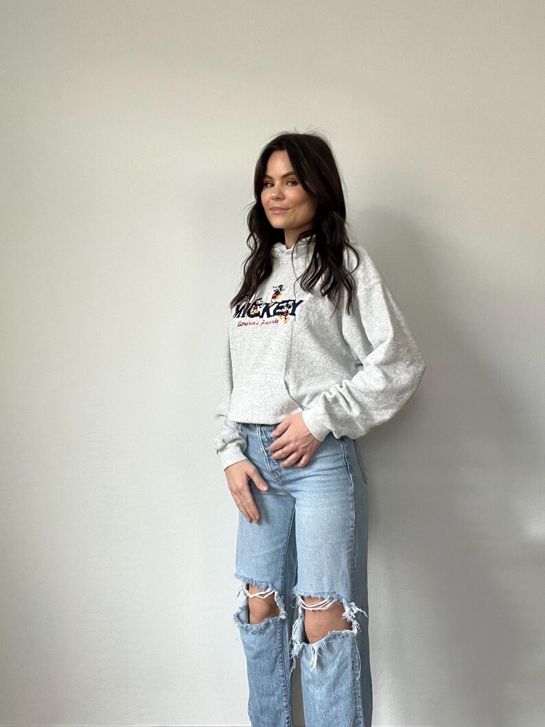 May include: A gray hooded sweatshirt with a Mickey Mouse graphic and the text "Mickey" and "Disney World" printed on the front. The model is wearing the sweatshirt with light blue ripped jeans.