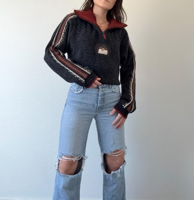 Puede incluir: Un su&eacute;ter gris oscuro con cuello rojo y cremallera de cuarto. Las mangas tienen detalles a rayas en crema, marr&oacute;n y rojo. El su&eacute;ter se combina con unos vaqueros rotos de color azul claro.