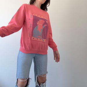 May include: A coral-coloured sweatshirt with a cat graphic. The cat is blue and grey, wearing a bow, set against a purple background with a border of hearts. The sleeves have a decorative design. The sweatshirt is a casual style.