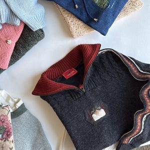 Puede incluir: Una variedad de su&eacute;teres doblados en varios colores, incluyendo azul, gris y rosa. Un su&eacute;ter gris oscuro presenta un cuello rojo y un peque&ntilde;o parche bordado. Tambi&eacute;n se ve una sudadera con capucha de mezclilla con un paisaje bordado.