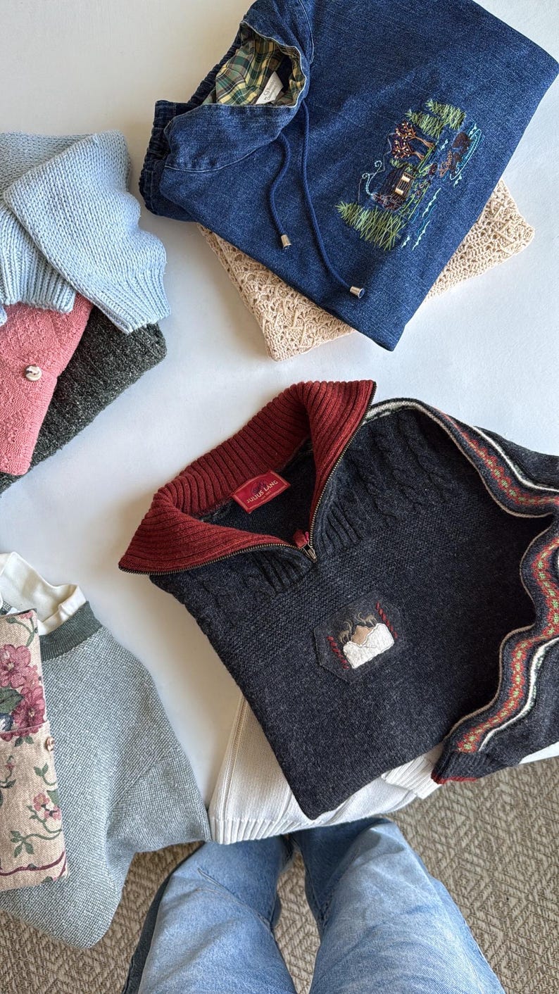 Puede incluir: Una variedad de su&eacute;teres doblados en varios colores, incluyendo azul, gris y rosa. Un su&eacute;ter gris oscuro presenta un cuello rojo y un peque&ntilde;o parche bordado. Tambi&eacute;n se ve una sudadera con capucha de mezclilla con un paisaje bordado.