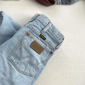 May include: Light blue Wrangler jeans with a brown leather patch and the Wrangler logo. A brown leather jacket and a blue and white sweater are also visible. The items are arranged on a white surface.