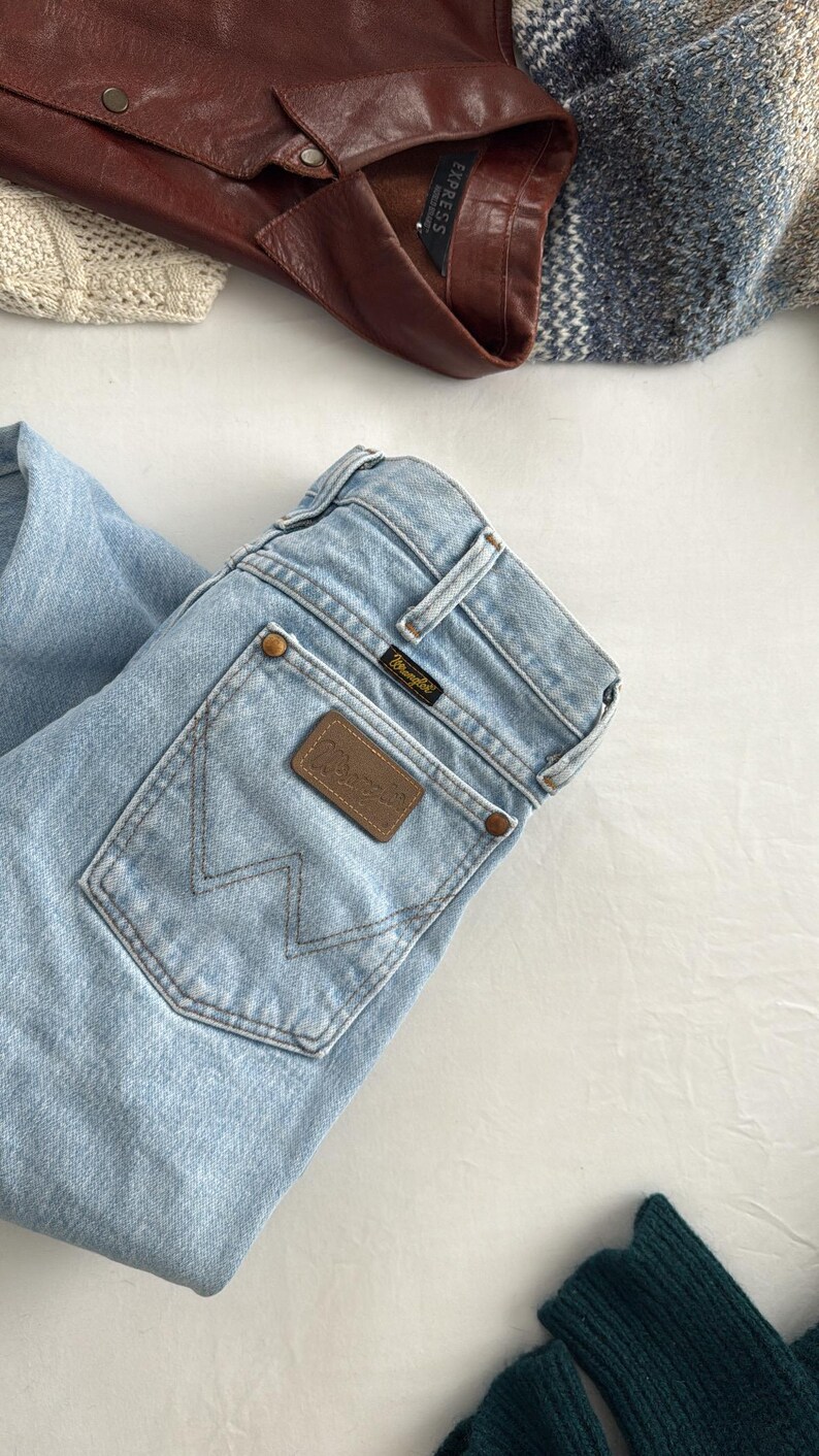May include: Light blue Wrangler jeans with a brown leather patch and the Wrangler logo. A brown leather jacket and a blue and white sweater are also visible. The items are arranged on a white surface.
