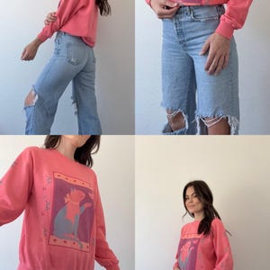 May include: Coral-coloured sweatshirt featuring a cat graphic wearing a scarf. The cat is within a rectangular frame with a purple background. Floral design on the sleeves. The sweatshirt is worn with light blue ripped jeans.