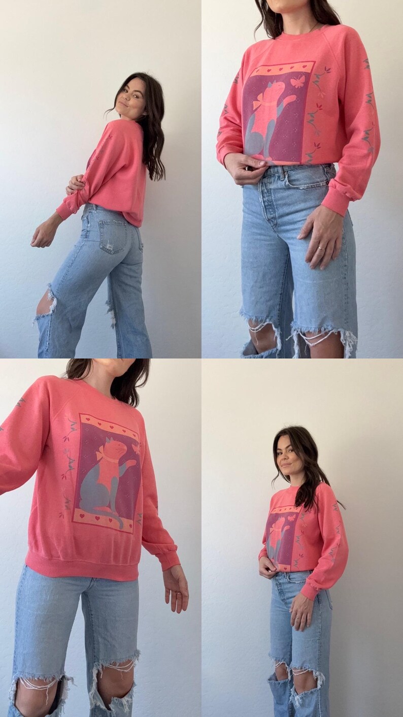 May include: Coral-coloured sweatshirt featuring a cat graphic wearing a scarf. The cat is within a rectangular frame with a purple background. Floral design on the sleeves. The sweatshirt is worn with light blue ripped jeans.
