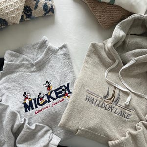 May include: Two gray sweatshirts with embroidered designs. One sweatshirt has a Mickey Mouse design with the text "Mickey America's Favorite". The other sweatshirt has a sailboat design with the text "Walloon Lake".