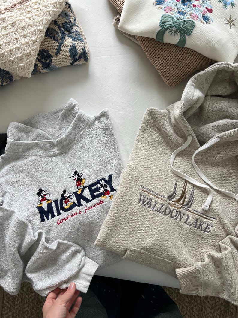 May include: Two gray sweatshirts with embroidered designs. One sweatshirt has a Mickey Mouse design with the text "Mickey America's Favorite". The other sweatshirt has a sailboat design with the text "Walloon Lake".
