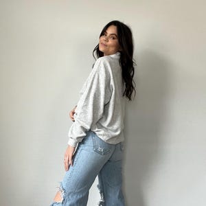 May include: A woman wearing a gray sweater and light blue jeans with distressed details. The jeans have a wide leg style.
