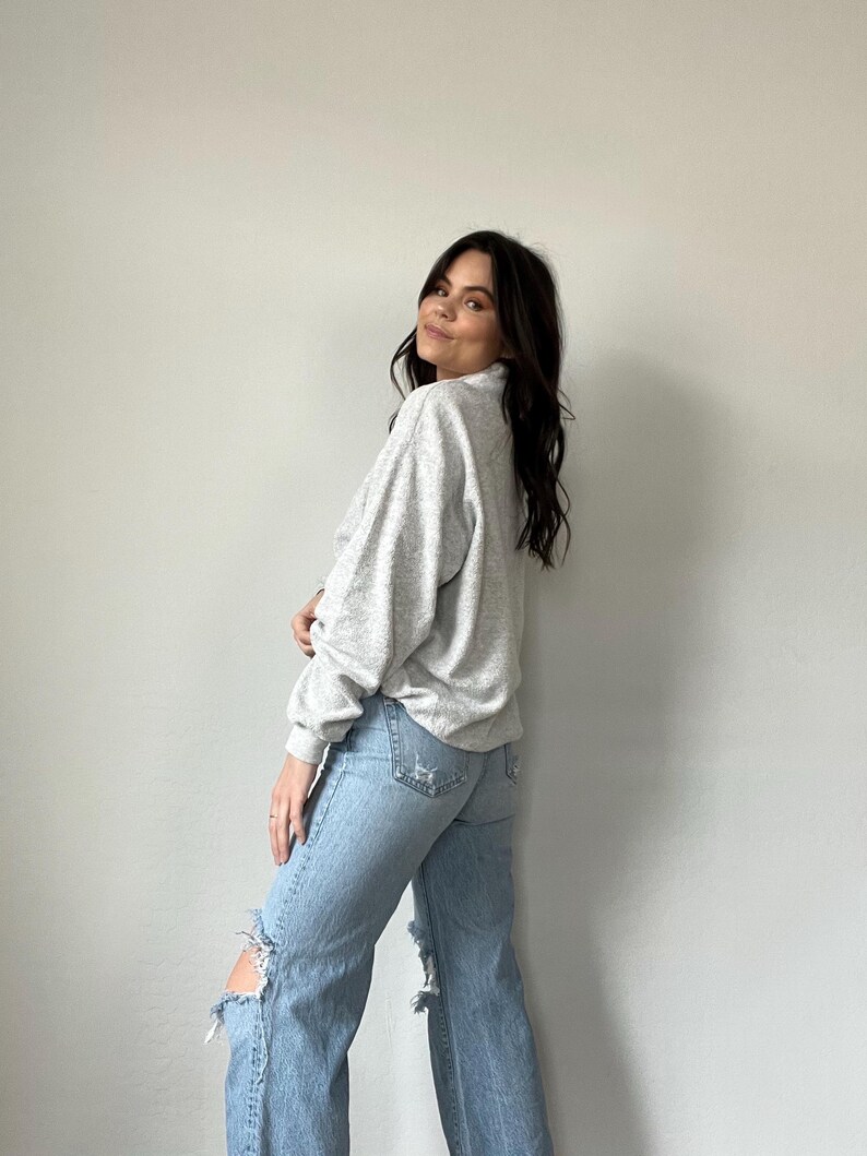 May include: A woman wearing a gray sweater and light blue jeans with distressed details. The jeans have a wide leg style.