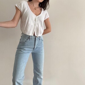 May include: A white, short-sleeved knit cardigan with a button-up front and a scalloped hemline. Paired with light blue denim jeans. The cardigan has a delicate, open-weave design. The jeans are high-waisted with a straight leg.